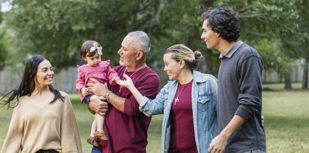 A multi-generation Hispanic family standing in the park together. The grandfather, a mature man in his 50s, is holding his granddaughter, a 16 month old baby girl. His adult daughter, the baby's mom, is on one side of him, and wife and their teenage son, 16 years old, is on the other side. They are all smiling and looking at the baby.