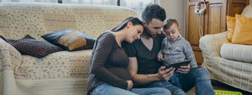 Family with small child and pregnant mother looking at the tablet sitting on the carpet